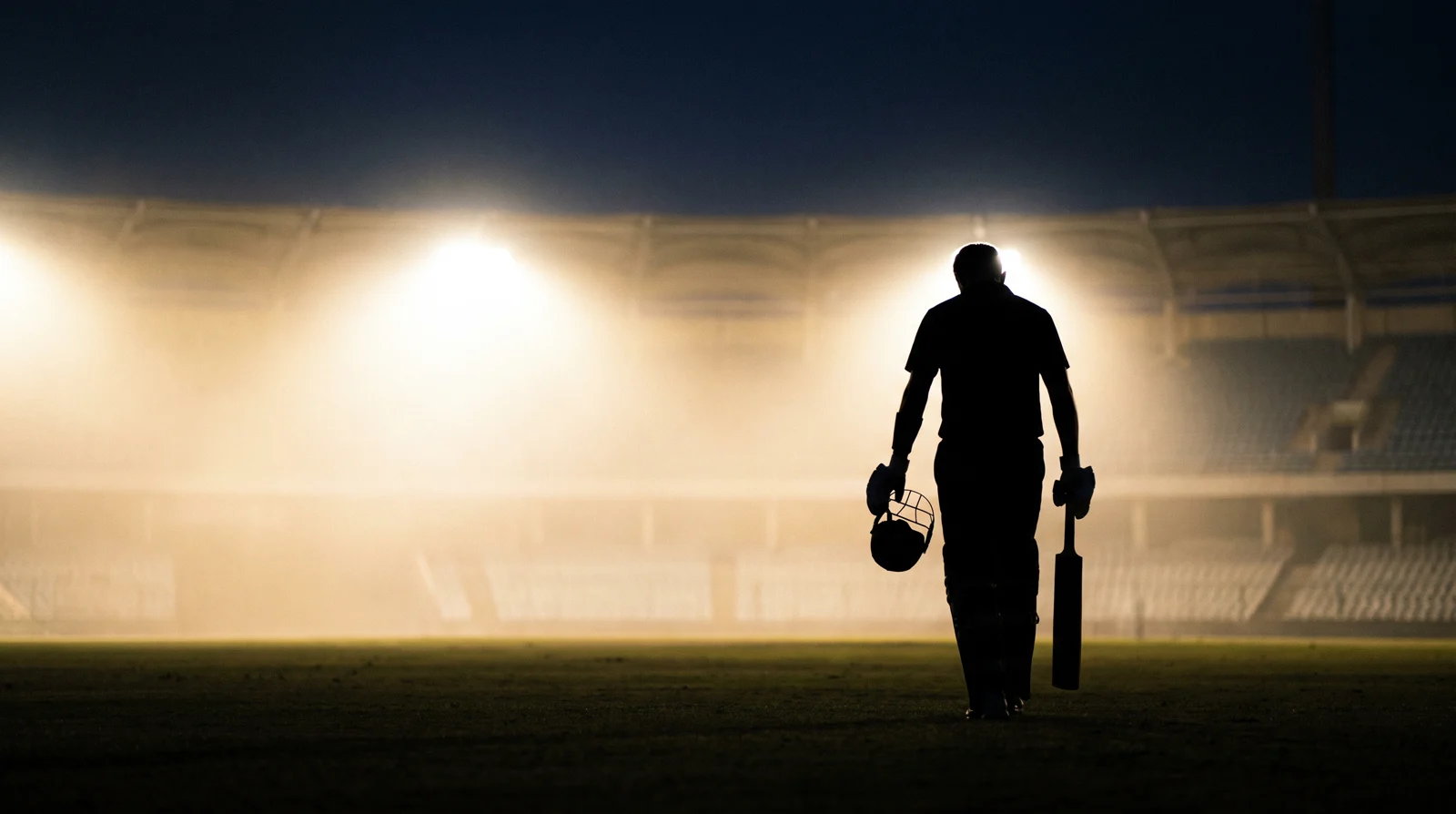 Silhouette of a batsman walking off the field alone at night, bat in one hand and helmet in the other, floodlit haze filling the empty stands behind him