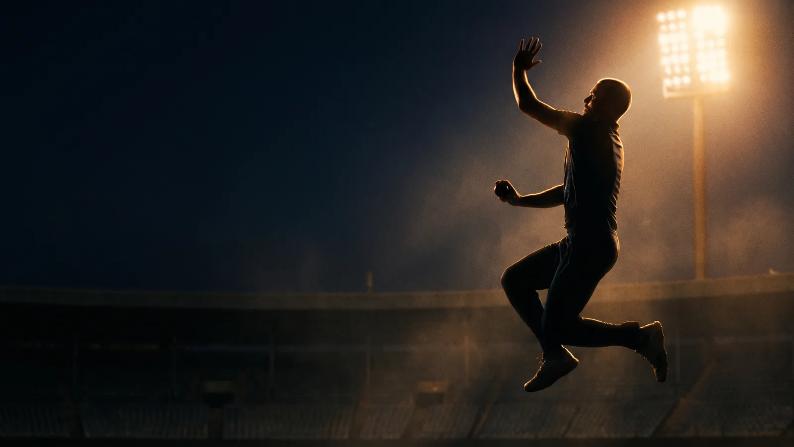 Silhouette of a bare-headed fast bowler frozen mid-leap in his delivery stride at night, rim-lit in amber by a single floodlight tower over empty dark stands